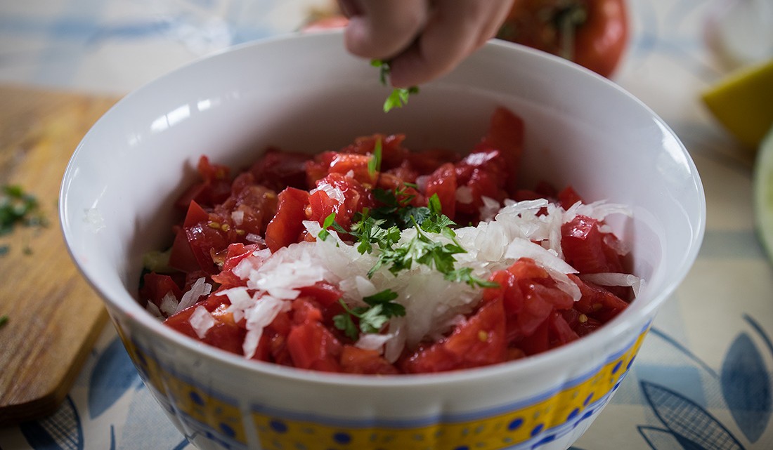Mão a espalhar salsa picada sobre salada israelita de pepino e tomate dentro de uma saladeira branca e dourada.