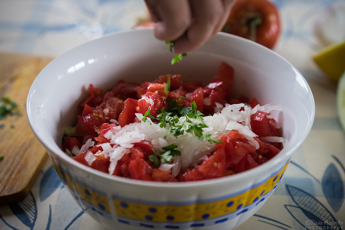 Mão a espalhar salsa picada sobre salada israelita de pepino e tomate dentro de uma saladeira branca e dourada.