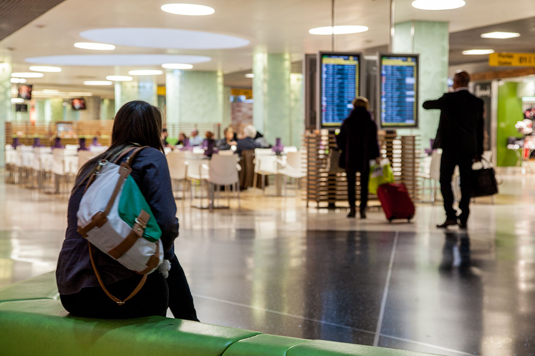 Viajante sentado num sofá e passageiros a olhar os painéis de informação do Aeroporto de Lisboa.