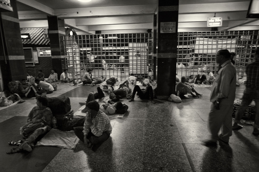 Várias pessoas sentadas ou deitas numa sala de espera na estação de comboios de Delhi.