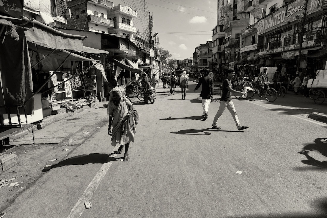 Rua de Varanasi com transeuntes e senhora idosa, envolvida em vários panos, a caminhar.