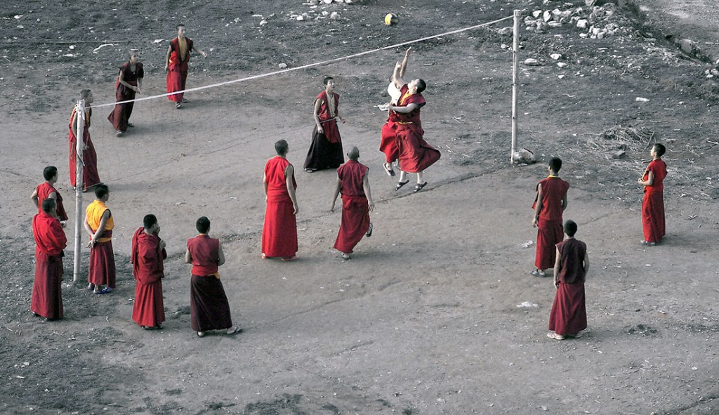 Monges budistas a jogar voleibol num terreiro com barreira feita de dois paus e uma corda.