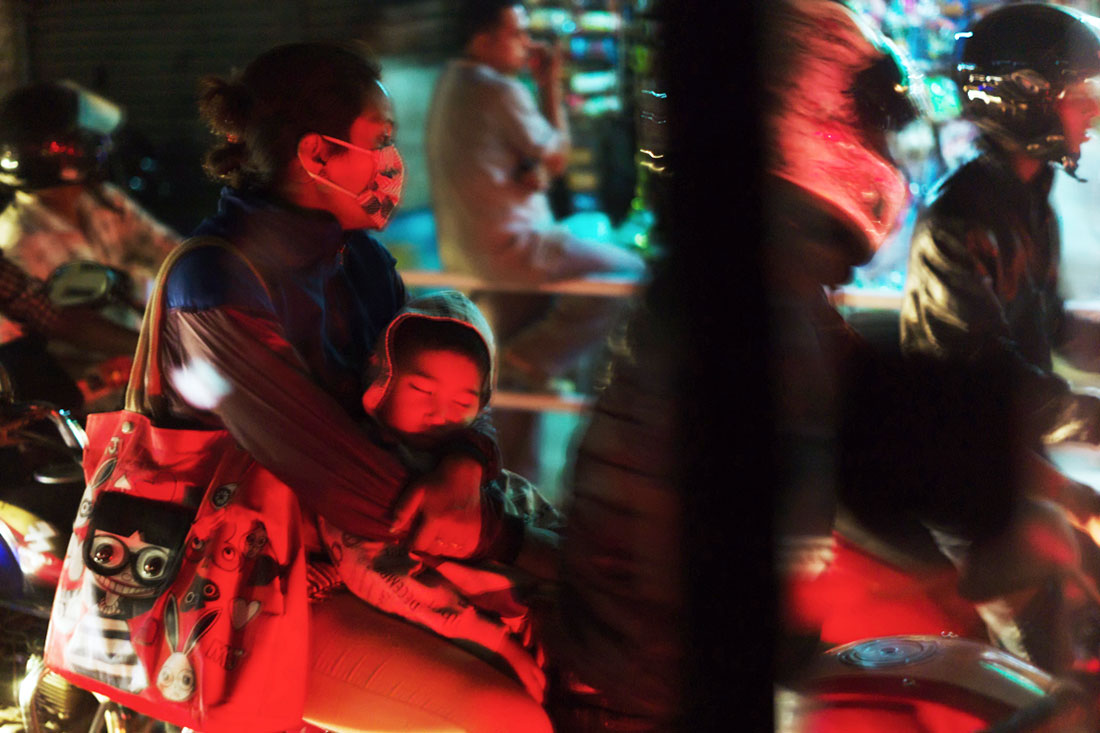 Mulher com criança adormecida ao colo, na traseira de uma mota e no meio do trânsito caótico de Kathmandu.