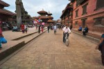 Homem de bicicleta e varias pessoas a caminhar na Praça Durbar em Kathmandu.