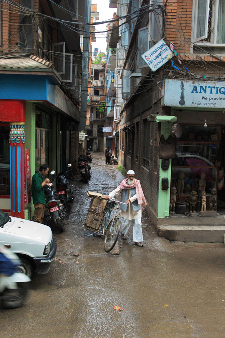 Homem conduz manualmente a sua bicicleta através de rua lamacenta em Kathmandu.