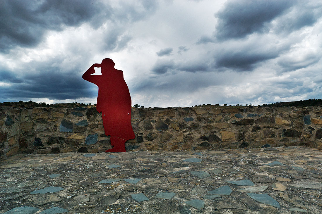 Silhueta vermelha de mouro junto à muralha de Mértola com céu carregado de nuvens.