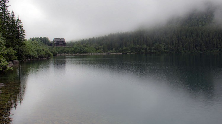 Espelho de água do lago Morskie Oko, envolto em nevoeiro, com casa abrigo ao fundo.